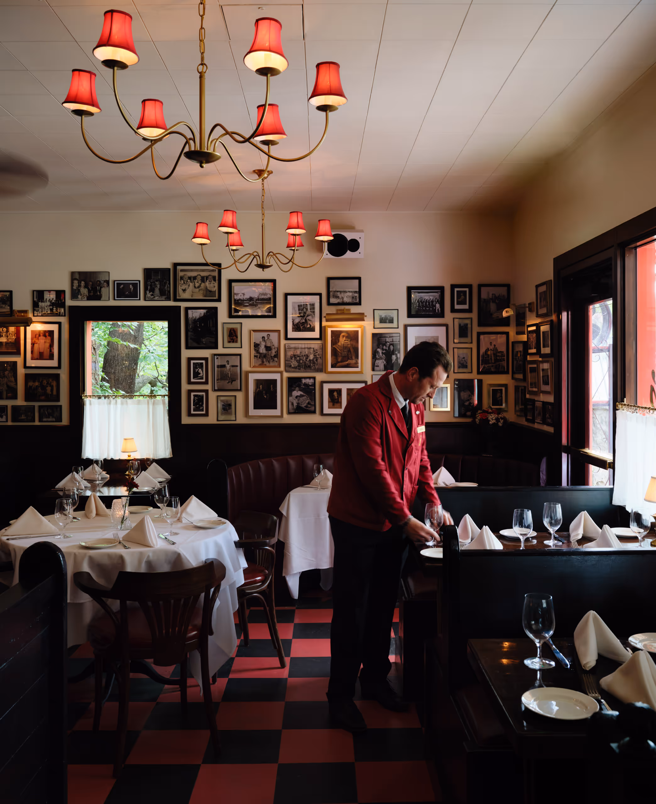 Server setting a dining table beneath red lamps and framed photographs.