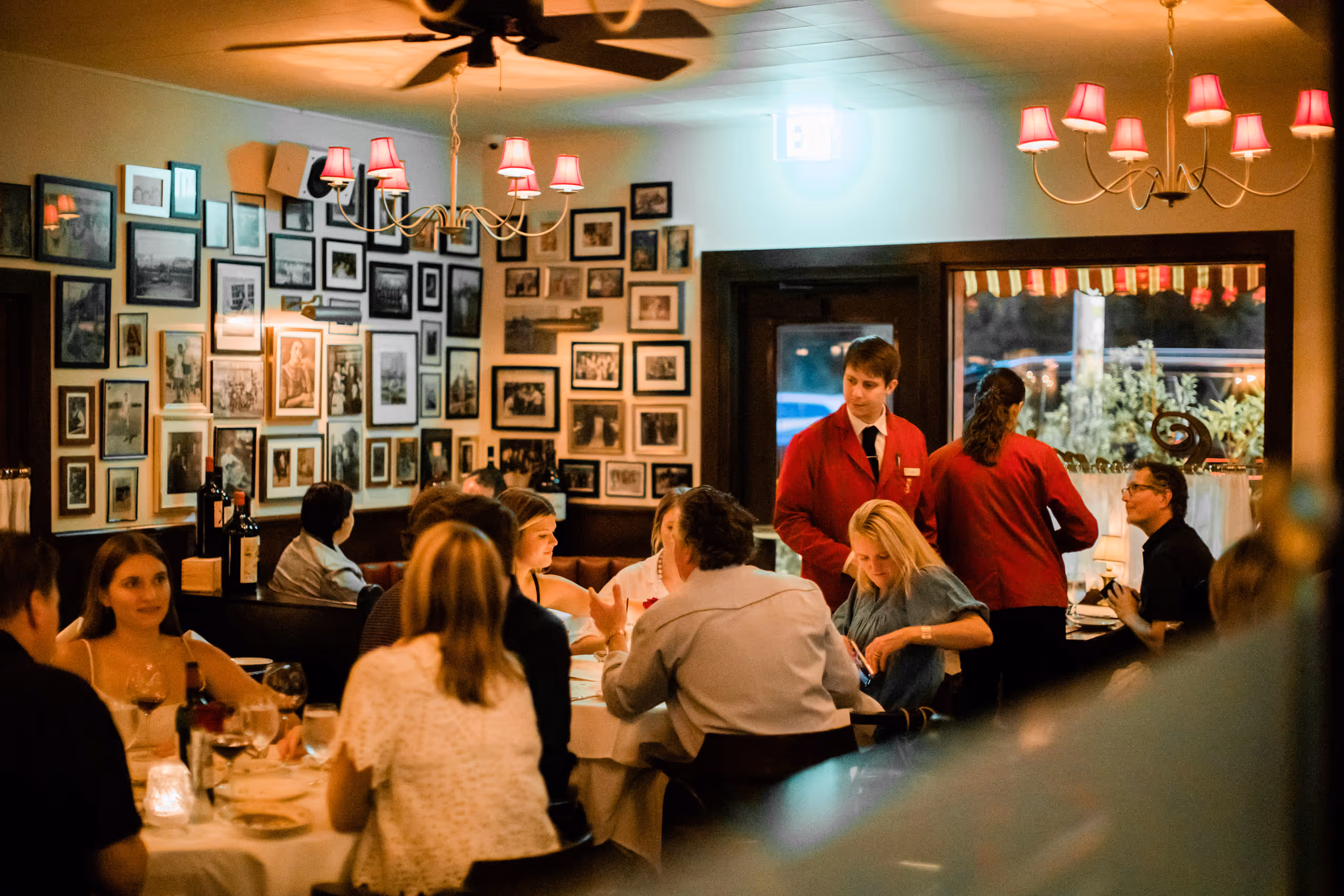 Guests and servers in the dining room beneath red lamps and framed photographs.