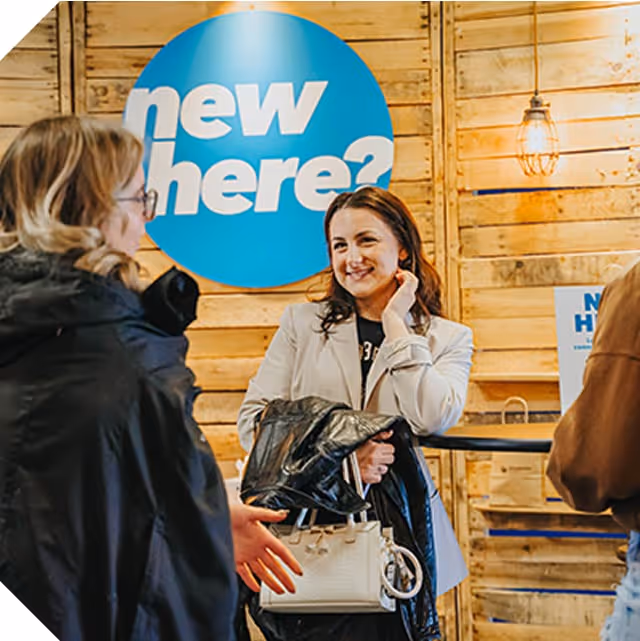 Two women talking together under a sign that says new here