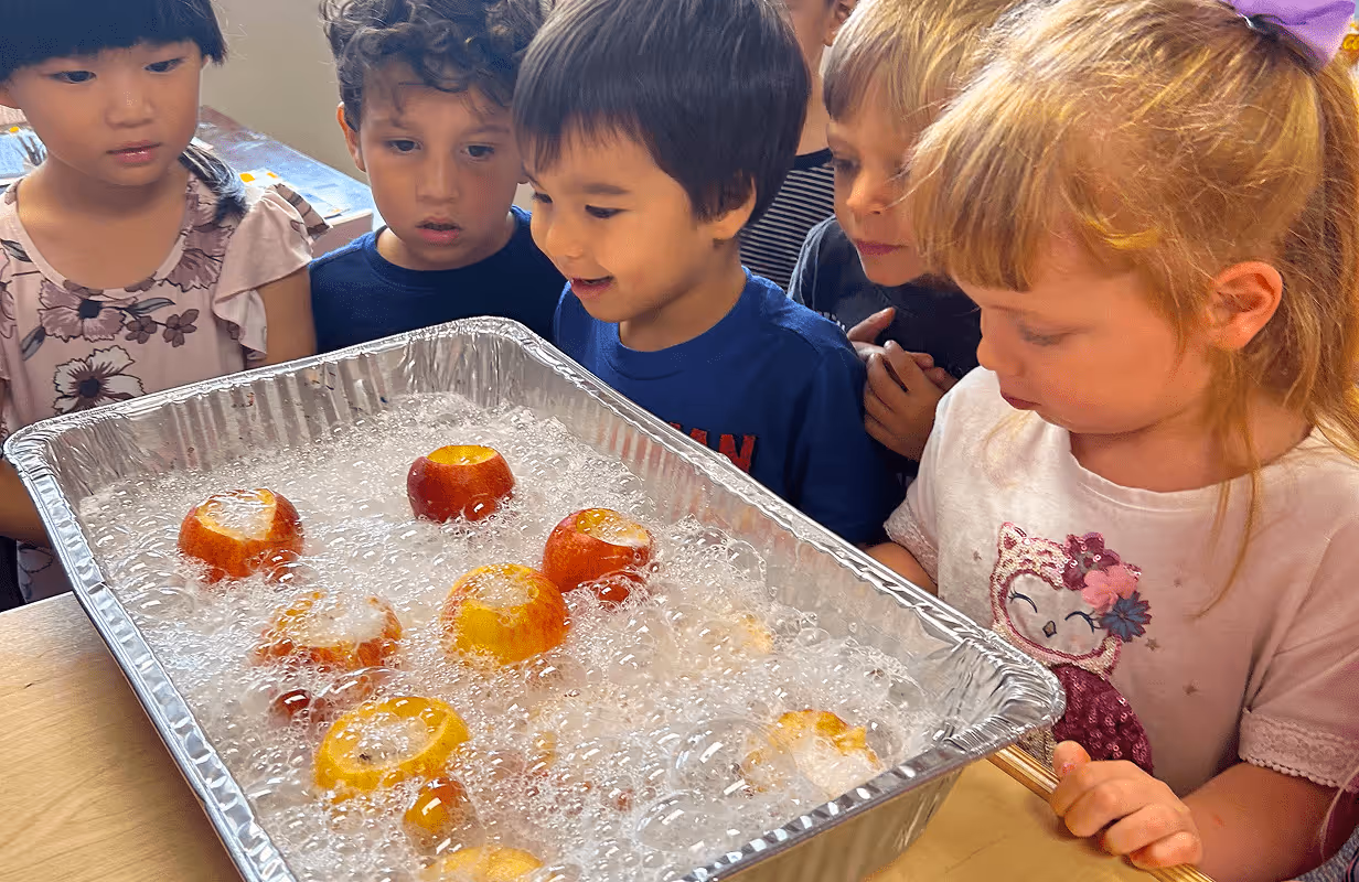 A group of young children gathered around a tray filled with apples and bubbles, watching with curiosity and excitement.