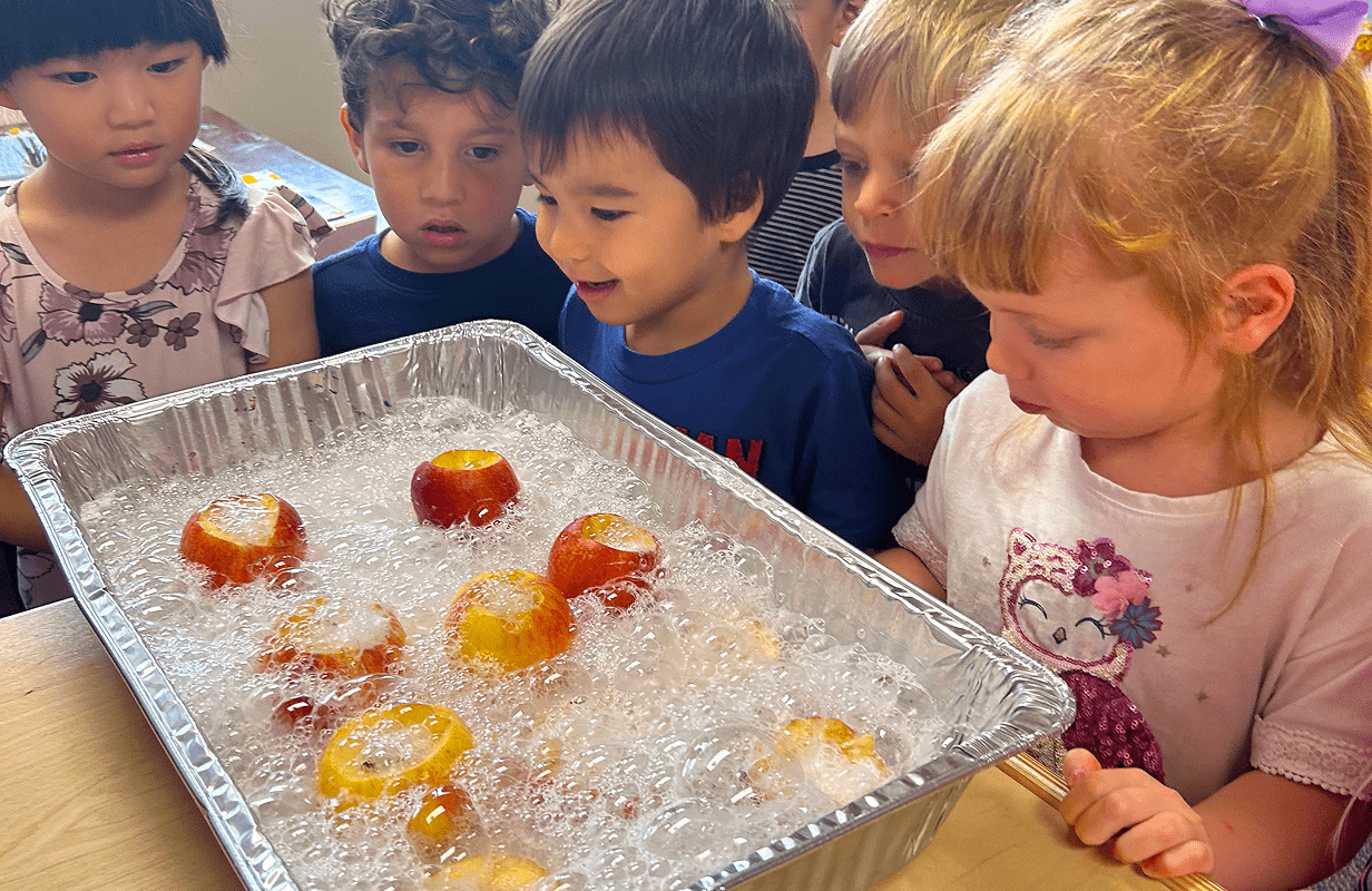 A group of young children gathered around a tray filled with apples and bubbles, watching with curiosity and excitement.