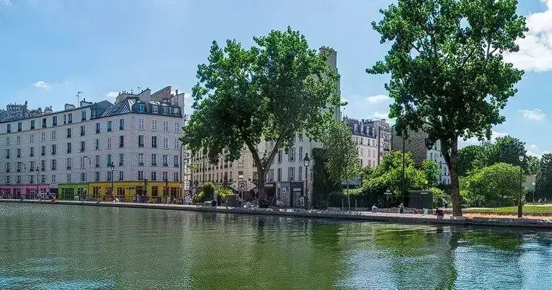 Bord de canal à Paris avec arbres verdoyants et bâtiments typiques, offrant un cadre charmant et paisible pour un appartement.
