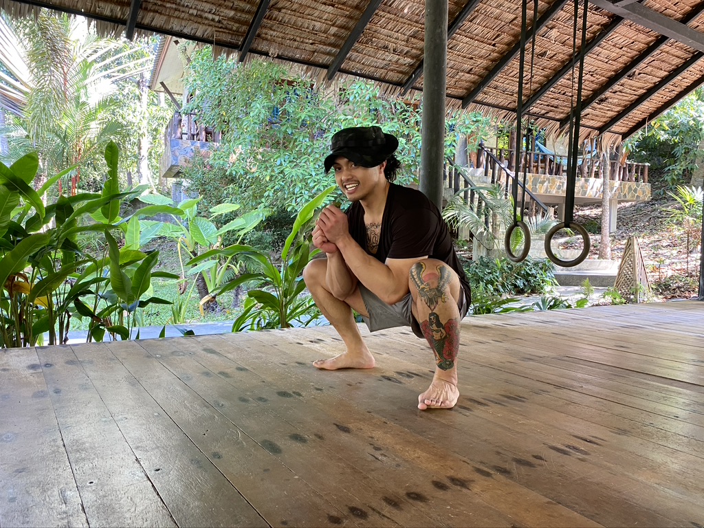 Man with a black hat and shorts squatting on a wooden floor under a thatched roof with green plants around.