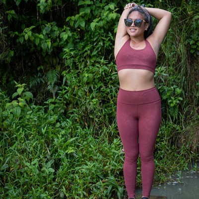 Woman in burgundy workout clothes stretching with hands behind head near dense green foliage.