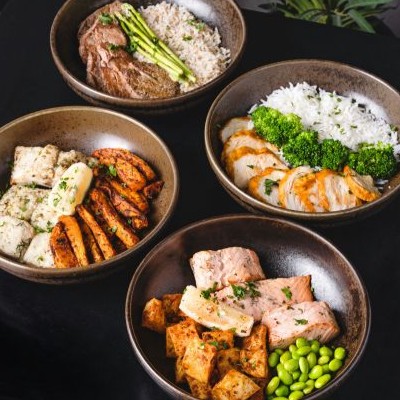 Four bowls with rice, grilled chicken or fish, roasted potatoes, edamame, and asparagus on black surface.