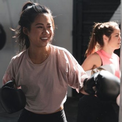 Two women smiling and boxing indoors, one in a light pink shirt and another in a pink tank top.