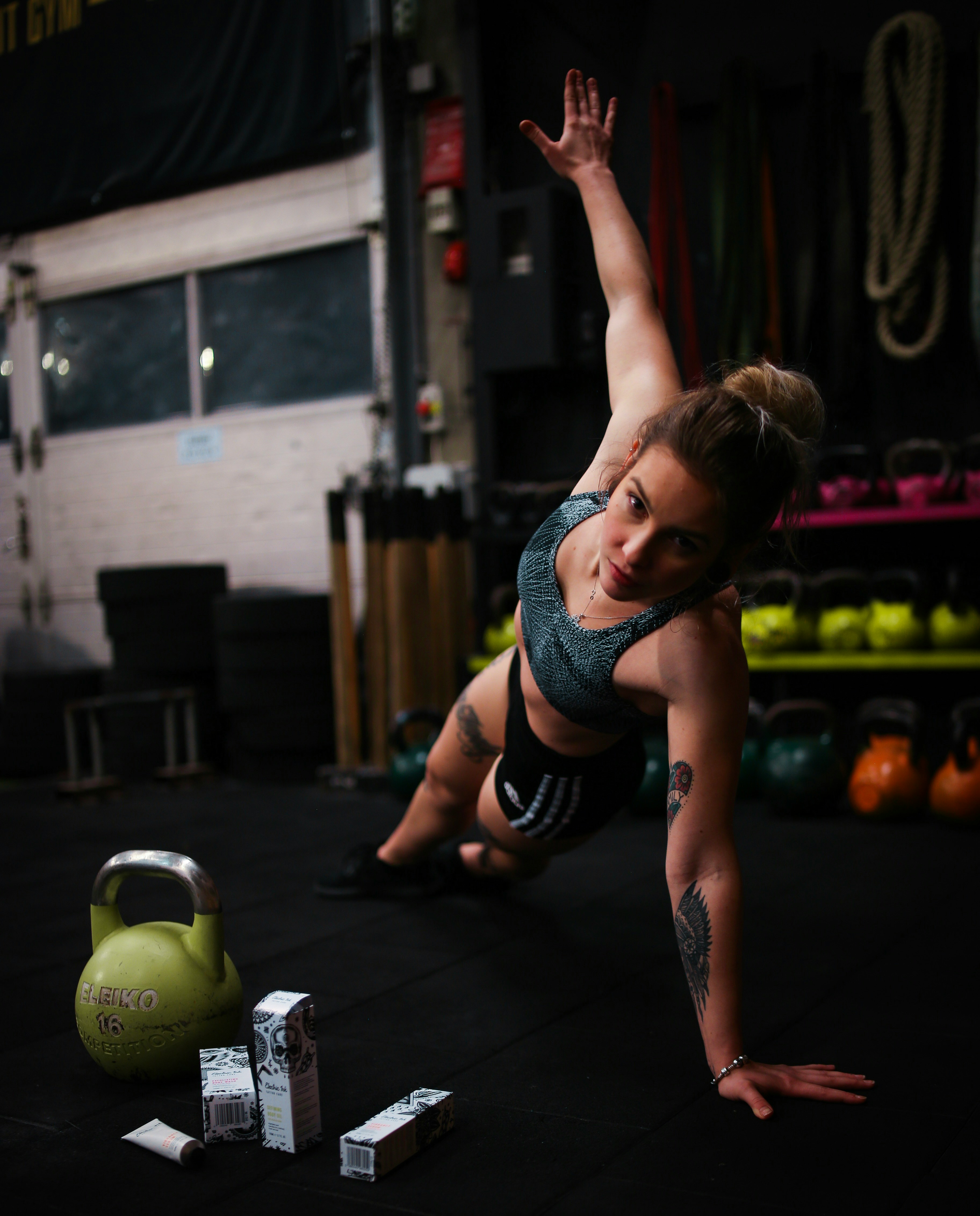 Woman with tattoos doing side plank exercise in gym near kettlebells and product boxes on floor.