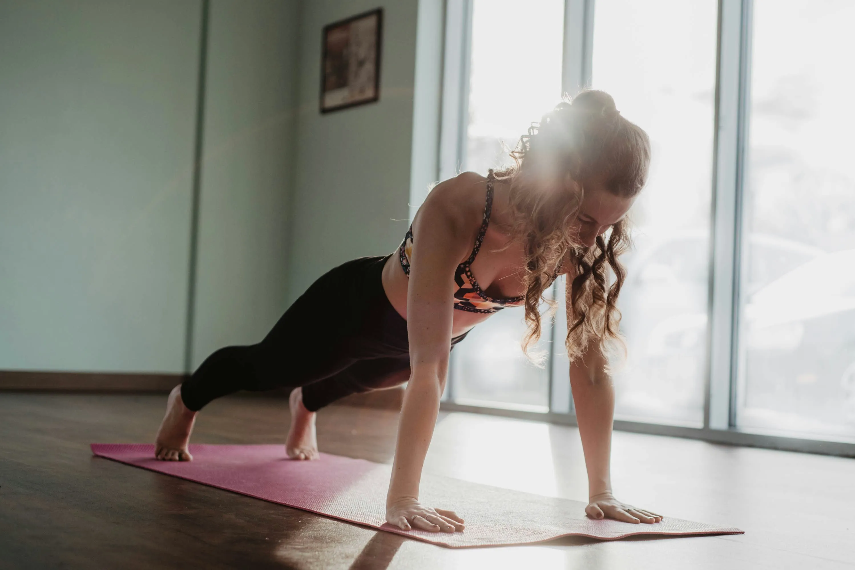 Woman doing a plank exercise on a pink yoga mat in a sunlit room with wooden floor.