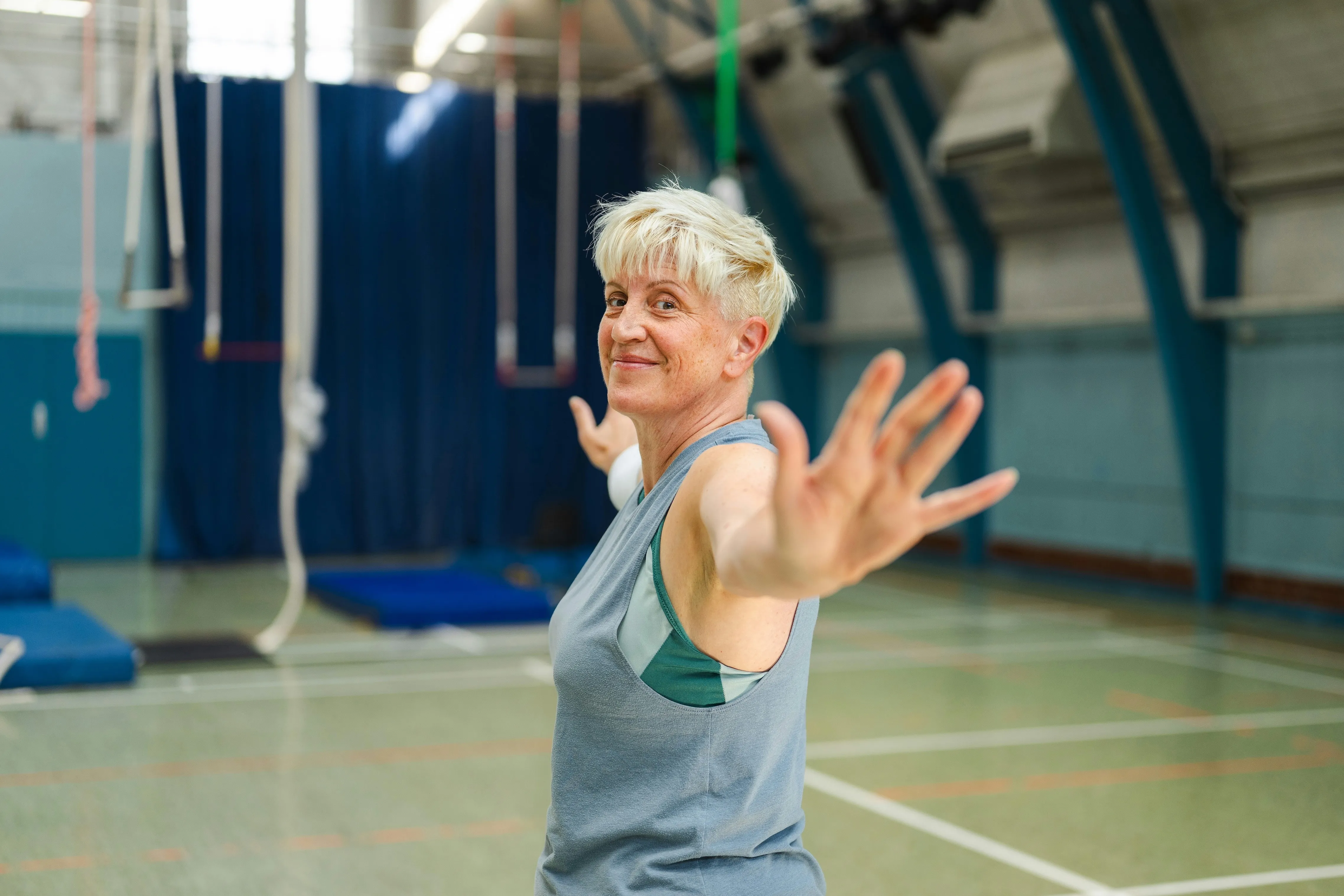 Mature woman in gym clothes does stretching or yoga pose with arms extended inside gymnasium.