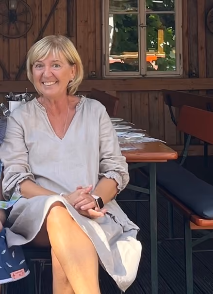 Lächelnde Frau mit kurzem blondem Haar und beigem Kleid sitzt in einer Holzgaststätte vor einem gedeckten Tisch.