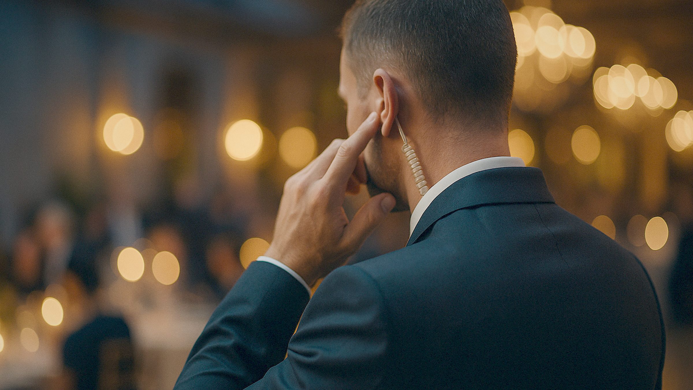 A close, elegant detail of a security team lead in a dark suit at an evening event, touching a discreet earpiece, a softly li