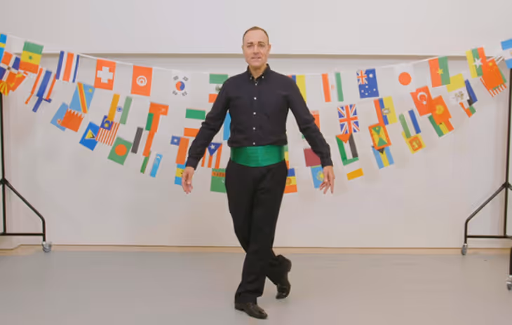 A performer in Irish dance attire, wearing a black outfit with a green sash, dances in front of a backdrop of international flags, symbolizing cultural diversity in our school dance workshops.

