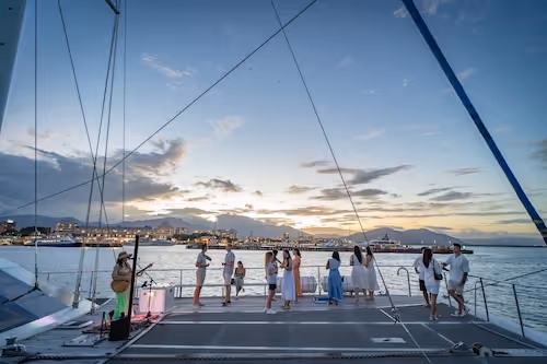 Group of people socializing on a large sailboat at sunset with city lights and mountains in the background.