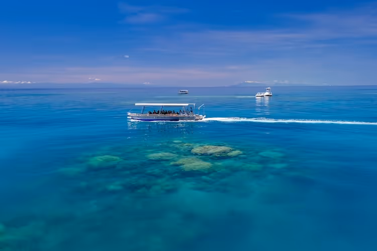 Boat with passengers cruising on clear blue ocean water above visible coral reef under a partly cloudy sky.