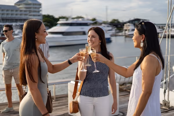 Three women smiling and toasting with champagne glasses on a dock with boats in the background.