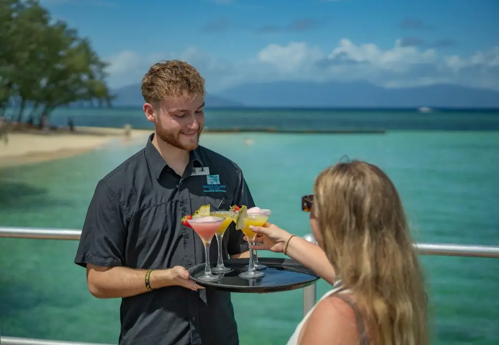 Smiling waiter serving colorful cocktails on a tray to a woman by the seaside with clear blue water and sky.