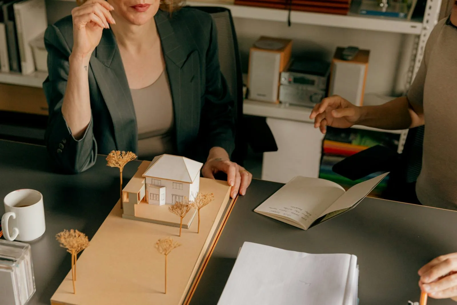 Two people discussing a small architectural model of a house on a table with a notebook, papers, and a coffee mug.