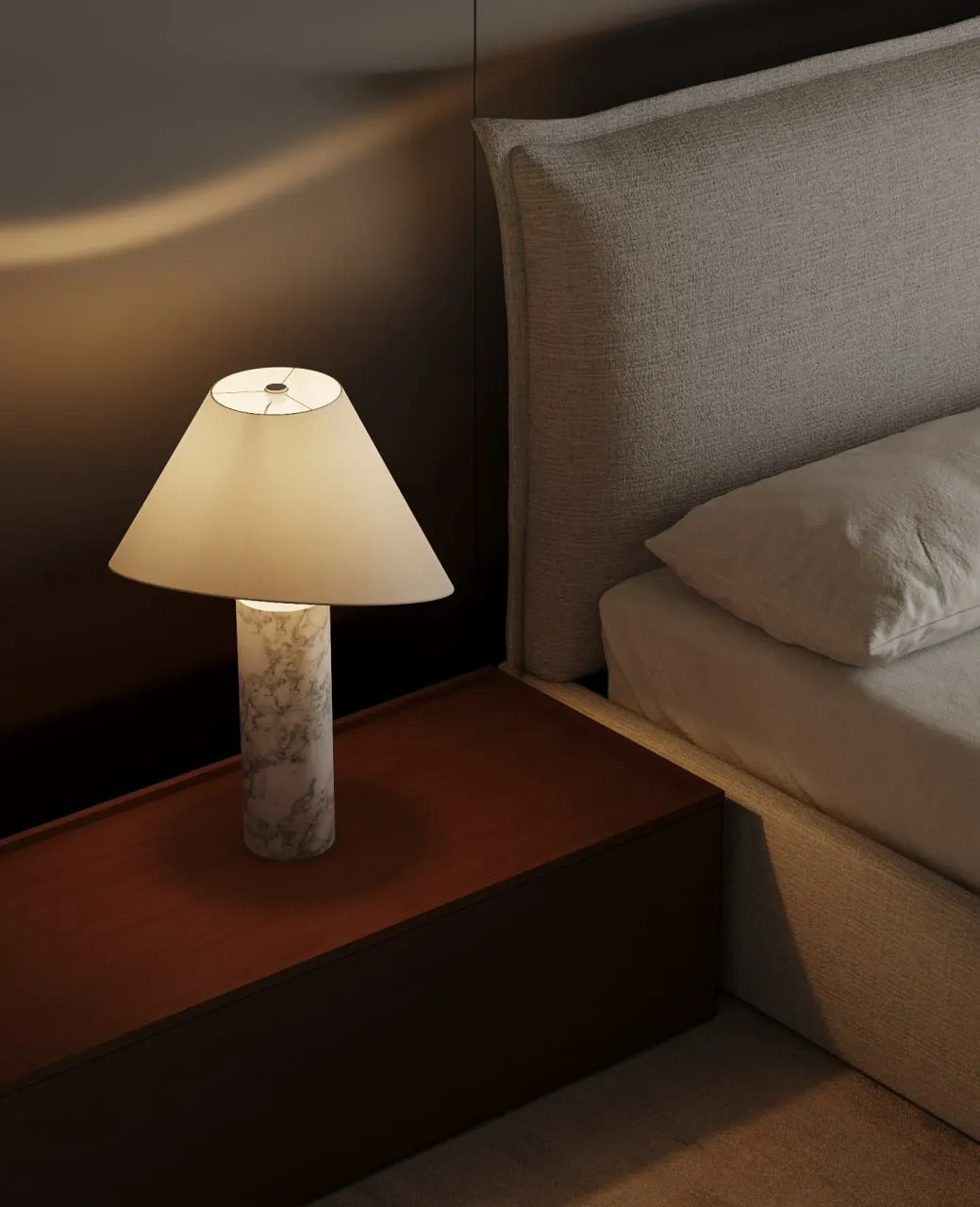 Lit table lamp with a marble base on a wooden nightstand beside a beige upholstered bed with a pillow.