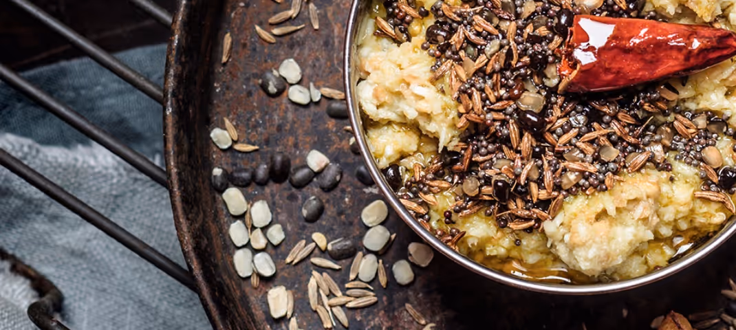 Close-up of a traditional Indian dish topped with tempered spices, red chili, and seeds, served in a metal bowl.