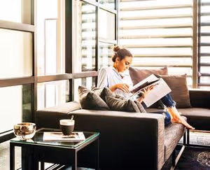 Woman sitting comfortably on a modern sofa in a sunlit room, reading a magazine with a coffee on the side table.