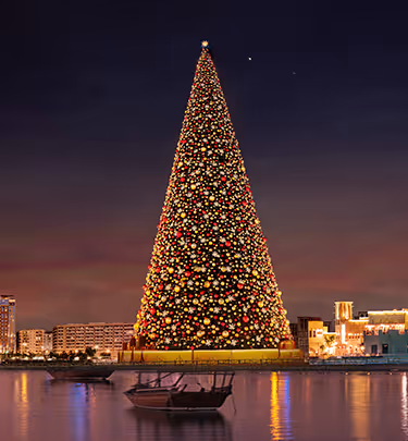 A giant illuminated Christmas tree by the waterfront at night, with reflections shimmering on the water and city buildings glowing in the background.  
