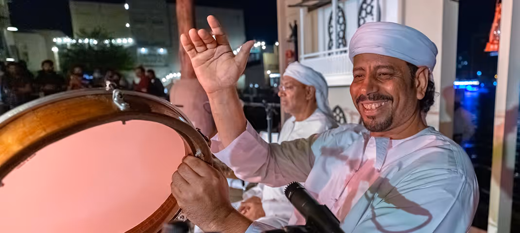Smiling man in traditional attire playing a large hand drum during a cultural performance at night.