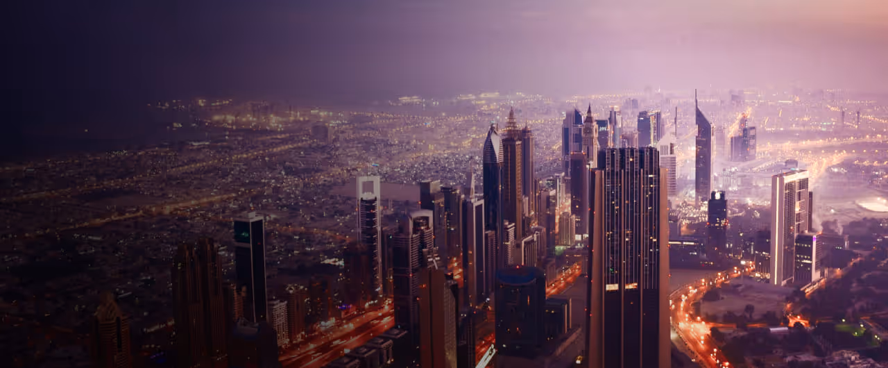 Aerial view of a modern city skyline at dusk, with towering skyscrapers illuminated against the hazy evening sky.