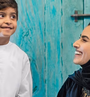 Smiling woman in an abaya looking at a cheerful young boy in traditional attire in front of a rustic turquoise door.