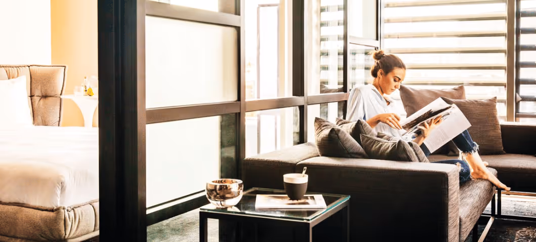 Woman relaxing and reading a magazine in a modern, sunlit hotel suite with a cozy seating area.