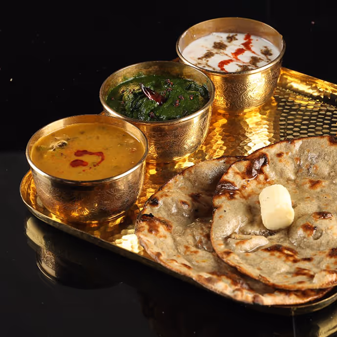 Traditional Indian meal served on a golden tray, featuring buttered parathas with dal, saag, and raita in brass bowls.