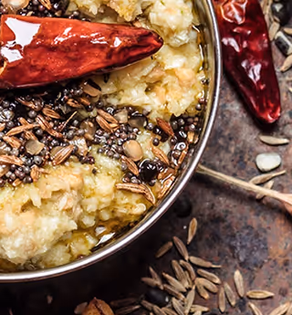 Close-up of a traditional Indian dish topped with tempered spices, red chili, and seeds, served in a metal bowl.