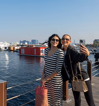 Two women smiling and taking a selfie by the waterfront with boats and city buildings in the background.