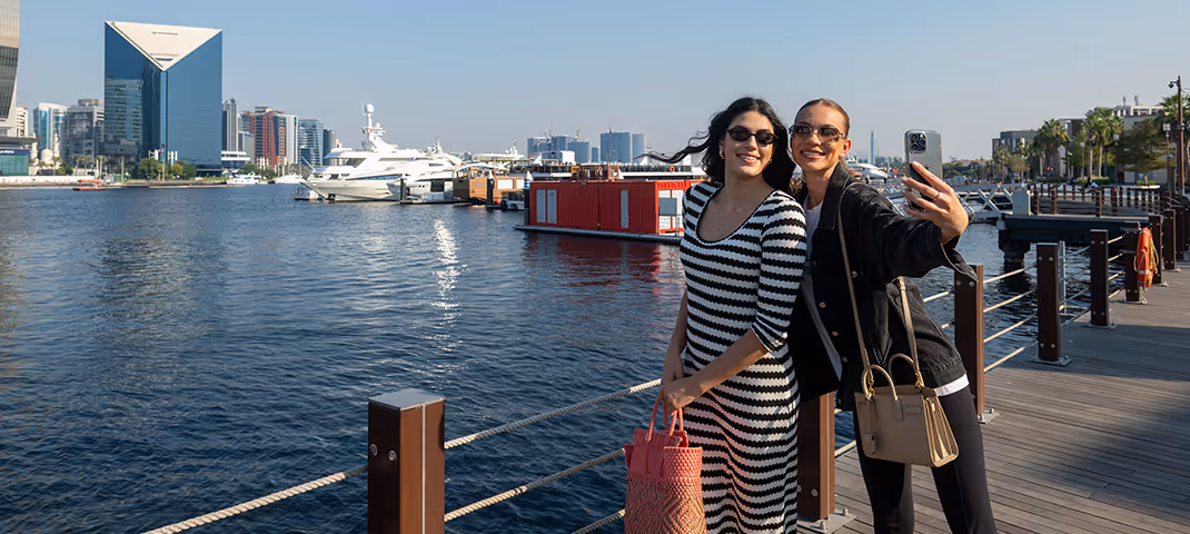 Two women taking a selfie by the waterfront with boats, city buildings, and the Dubai Creek in the background.  
