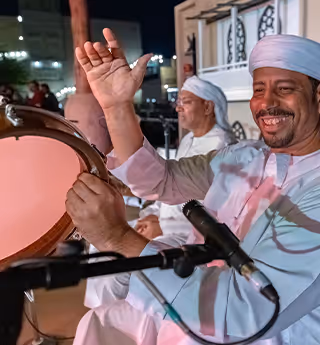 Man in traditional clothing smiling and playing a drum during a cultural performance, with other musicians in the background.  
