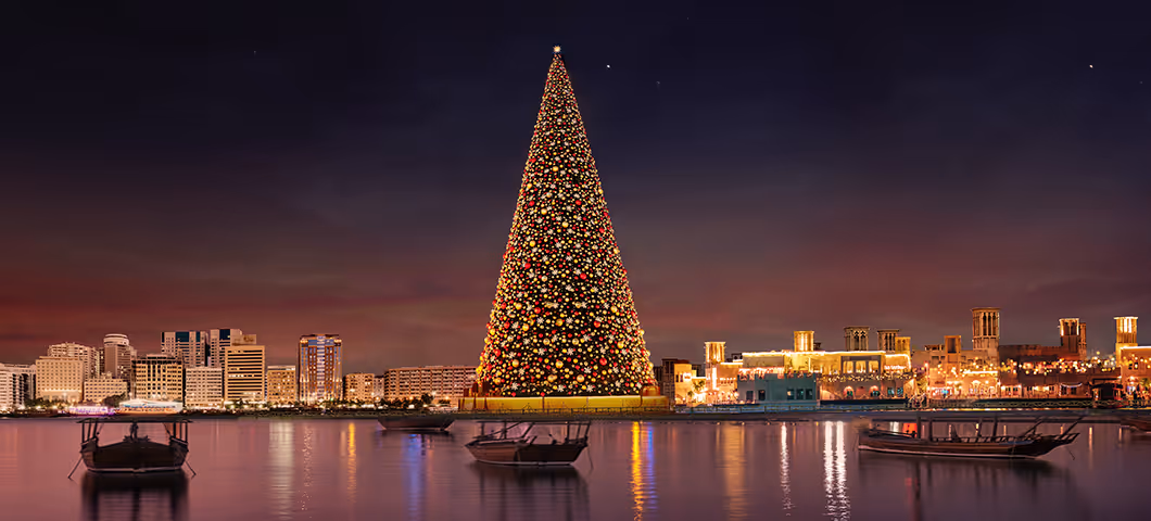 A giant illuminated Christmas tree by the waterfront at night, with reflections shimmering on the water and city buildings glowing in the background.  
