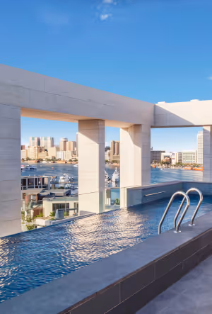 Rooftop infinity pool with cityscape views in the background, surrounded by modern white stone columns under a clear blue sky.