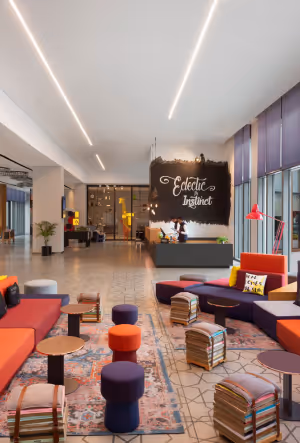 Modern hotel lobby with colorful seating, patterned rugs, low stools, and a welcoming reception desk under a chalkboard-style sign.