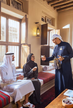 Traditional Arabic hospitality scene with a man in a kandura pouring coffee for a seated man and woman in traditional attire inside a sunlit majlis with wooden interiors.