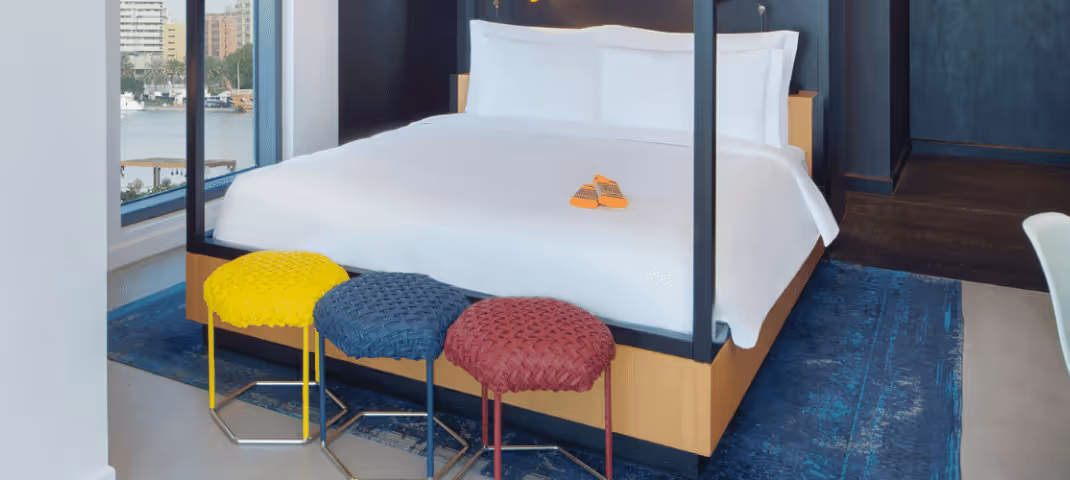 Modern hotel room with a large white bed, blue rug, and three colorful round stools at the foot of the bed.  
