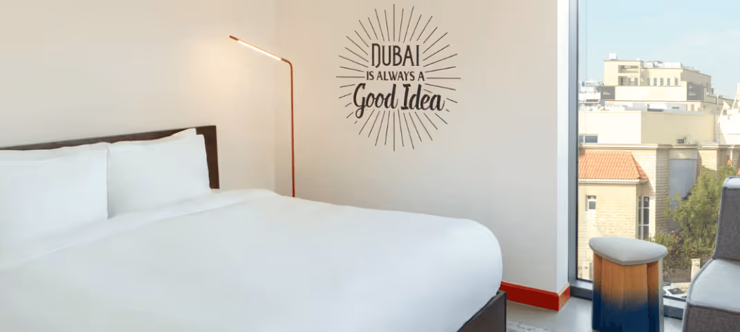 Minimalist hotel room with a neatly made bed, wall art reading "Dubai makes good ideas," and a window overlooking city buildings.