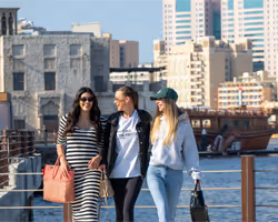 Three women walking outdoors by the waterfront, carrying shopping bags, with tall city buildings in the background.  
