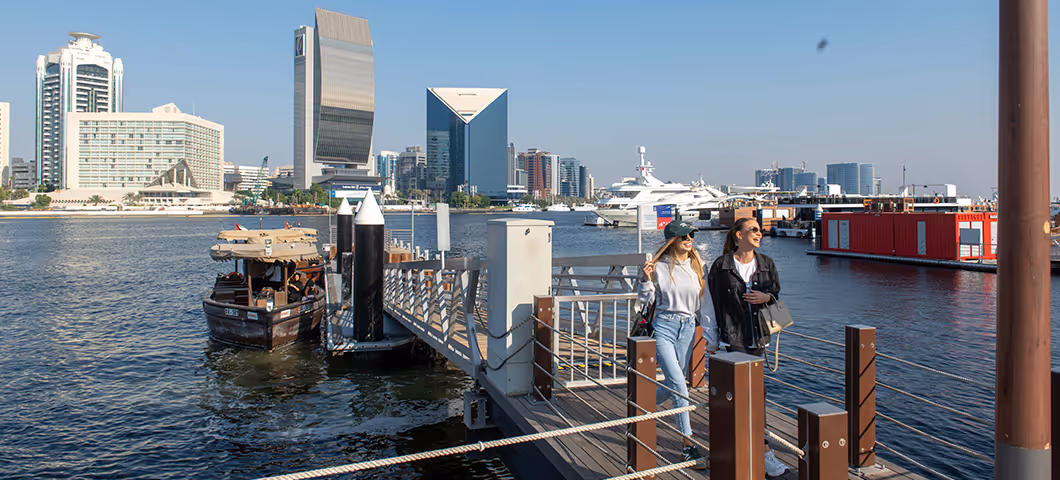 Three women enjoy drinks and conversation at an outdoor waterfront café at sunset, with lanterns overhead and boats visible on the water in the background.