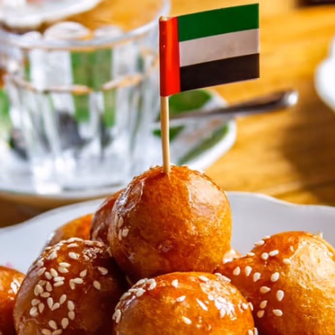 Close-up of golden luqaimat (fried dough balls) drizzled with syrup and sprinkled with sesame seeds, served with a small UAE flag on top.