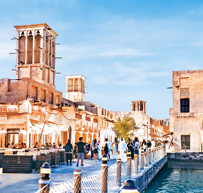 Crowds strolling through a traditional souq-like area with historic wind towers and Emirati-style buildings, near the water.
