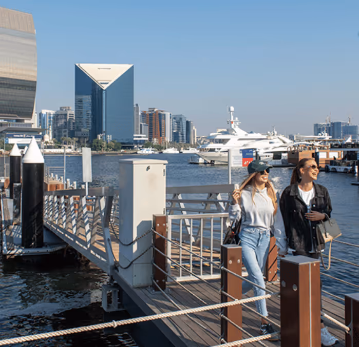 Two women walking along a marina boardwalk with yachts docked nearby and the modern Dubai skyline visible in the background.