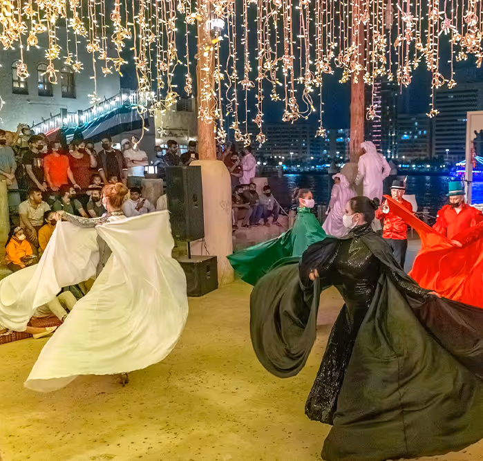 Colorfully dressed performers swirling their garments during a traditional dance performance under string lights, with a waterfront audience in the background.