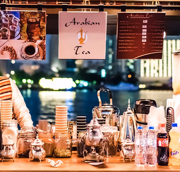 A traditional Arabian tea stall set up along the waterfront at night, displaying silver teapots, jars of herbs, and drink options under signs that read "Arabian Tea".