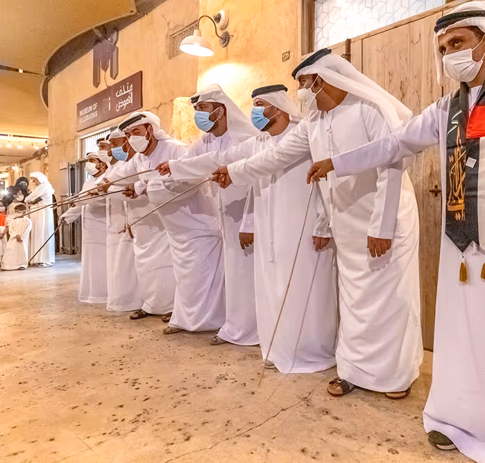 Group of Emirati men in white kanduras performing the Al Ayala stick dance outside a heritage building, wearing masks and standing in synchronized formation.