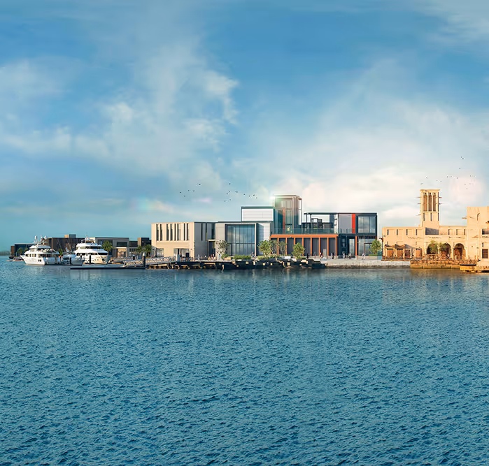 Scenic waterfront view featuring modern glass architecture beside traditional Emirati buildings with wind towers, under a clear blue sky.