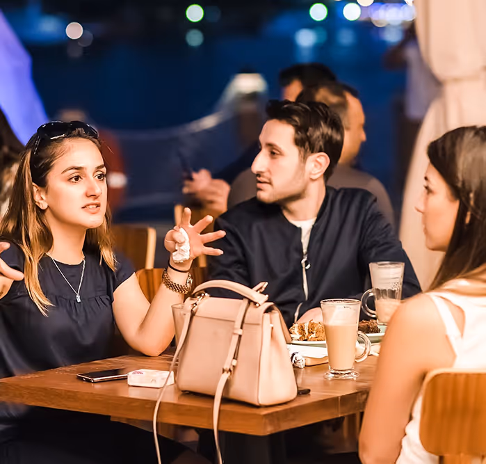 Group of young adults enjoying food and conversation at a waterfront restaurant, with city lights reflecting on the water in the background.